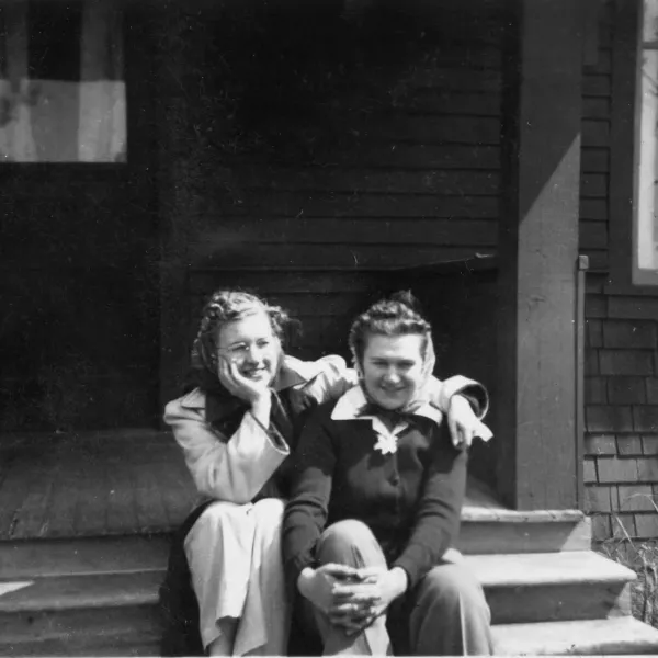 Two women sit next to each other smiling on the porch steps of Viqueen Lodge in 1940