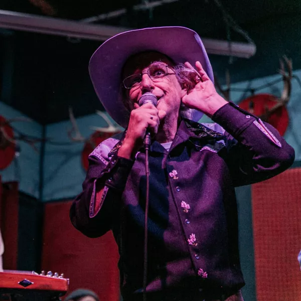 Patrick Haggerty, wearing a black cowboy shirt and lavender cowboy hat, sings on stage, holding one hand to his ear while he sings to encourage the audience to sing along. 