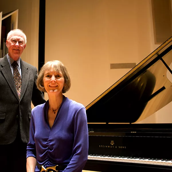 Ford Hill stands and Sybil Sanford is seated at a piano bench on stage in the WWU Concert hall