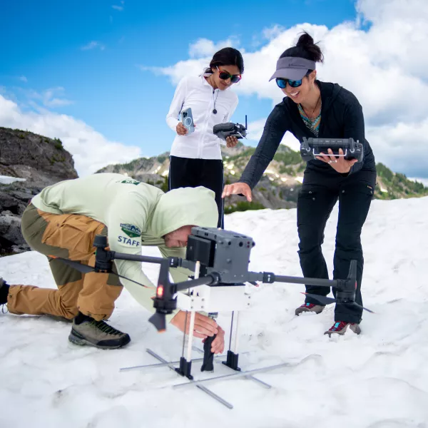 A student crouches down to adjust a piece of equipment on a drone parked on a snow bank on top of a mountain. Two other people are nearby with equipment and watch him work. 
