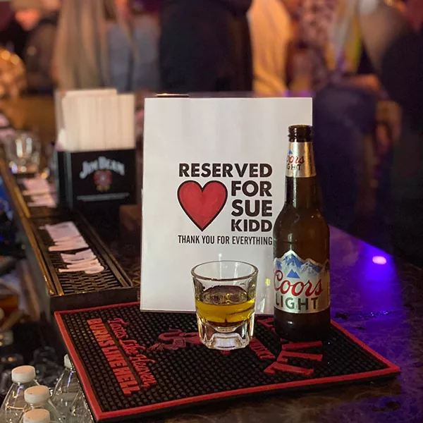 a bottle of beer and a shot of whiskey sit next to an empty chair at a crowded bar with a sign that says Reserved for Sue Kidd Thank You for Everything. 