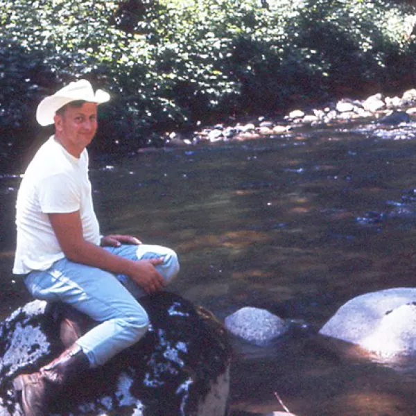 a man smiles, wearing a cowboy hat, white T, jeans and boots, sitting on a rock next to a stream. 