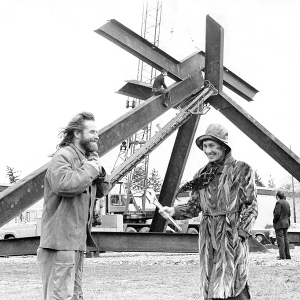 Mark di Suvero and Virginia Wright stand smiling and talking to each other in front of the sculpture For Handel. in 1974. Wright, wearing a stylish 70s-era fur trench coat and floppy rain hat, is holding a wrench.