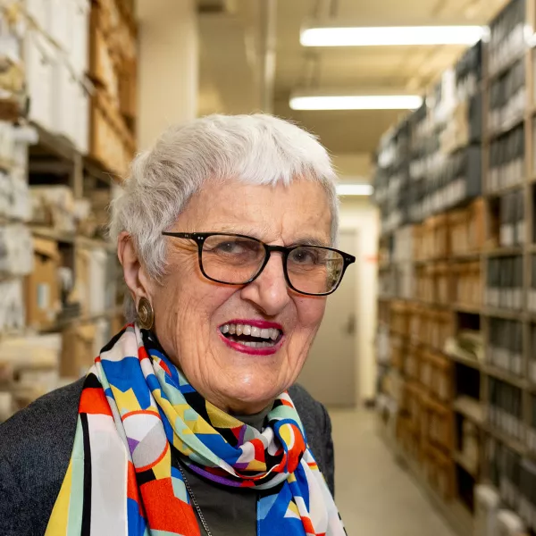 Catharine Stimpson smiles for the camera standing among the rows of books, boxes and scrolls in the archive storage room. 