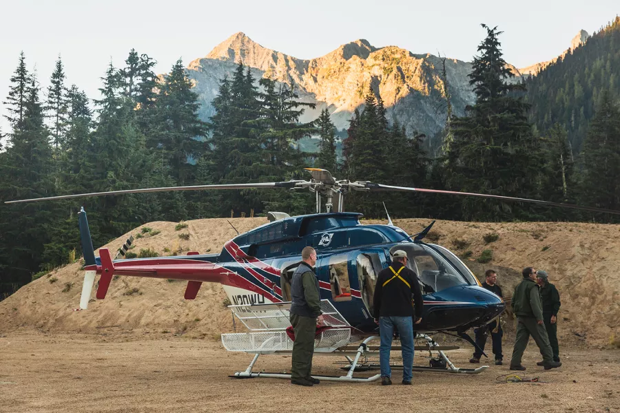 A helicopter sits idle with a view of tall, sunlit mountains in the background. 