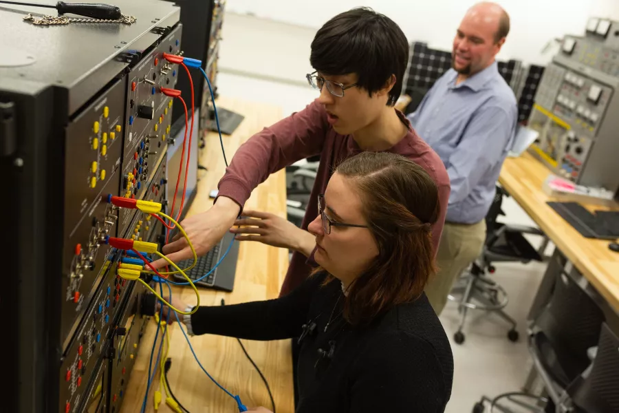 Two students adjust electronic equipment while their professor watches in the background. 
