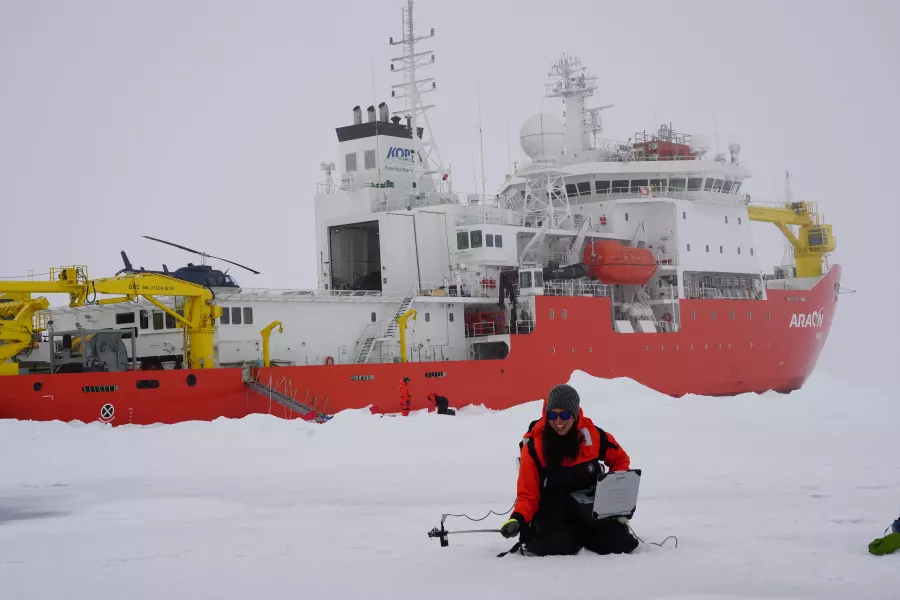 A researcher bundled for sub-zero temperatures sits on the ice in front of a orange icebreaker ship