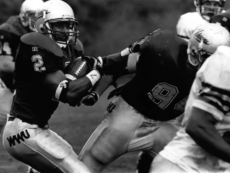 Steinauer in a WWU football uniform clutches a football in both arms and runs toward a group of other playersr