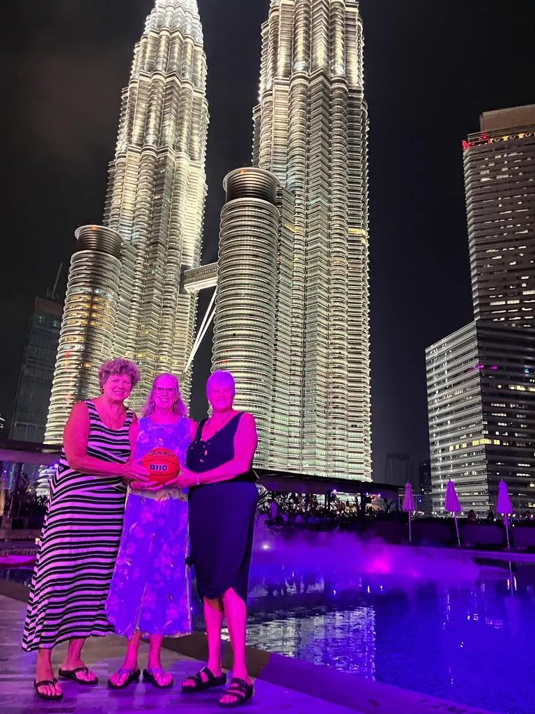 Three women hold a basketball in front of a soaring double skyscraper in Kuala Lumpur