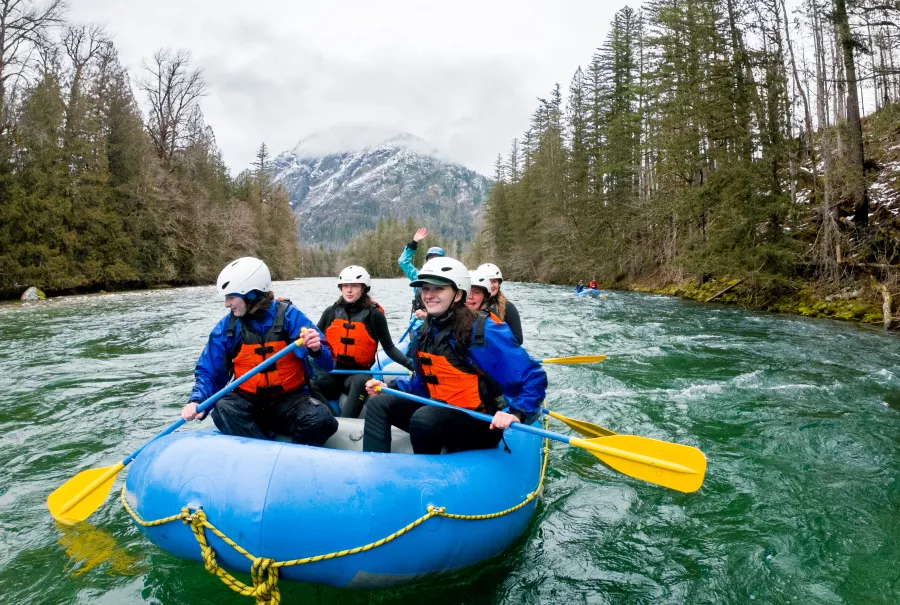 Six students paddle a raft into some minor rapids, a mountain peak dotted with snow in the background.. One smiles for the camera. 