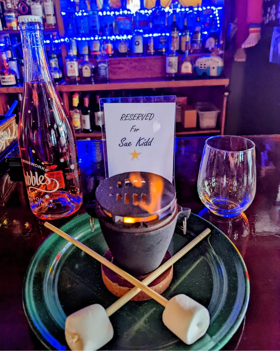 A plate with two sticks with fluffy marshmallows at the ends sit crossed next to a pot with a little flame. Nearby sit a small bottle of rose sparkling wine and a sign, Reserved for Sue Kidd.