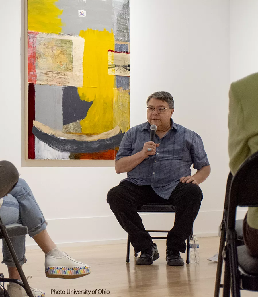 John Feodorov sits in front of a painting and speaks into a microphone to a gallery of seated listeners. 