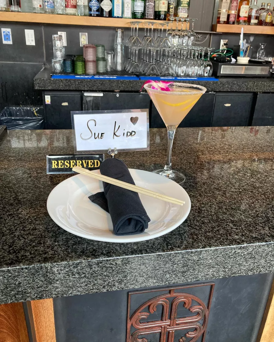 A fancy pink cocktail in a martini glass with an orange peel twist, a salt rim and a dainty orchid sit next to a Reserved sign with the name Sue Kidd