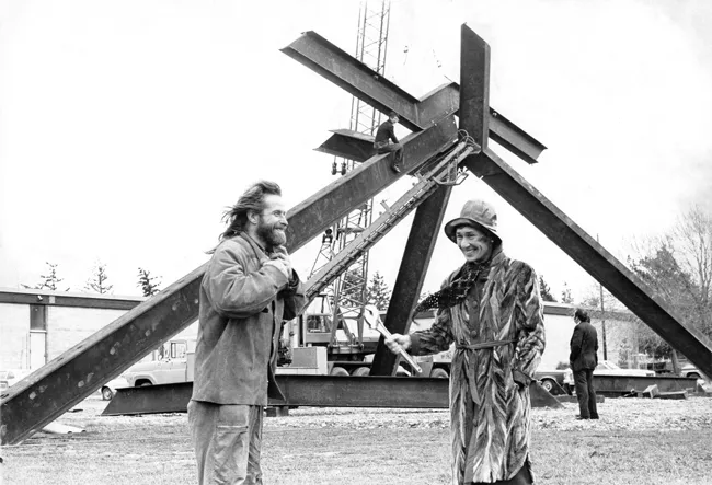 Mark di Suvero and Virginia Wright stand smiling and talking to each other in front of the sculpture For Handel. in 1974. Wright, wearing a stylish 70s-era fur trench coat and floppy rain hat, is holding a wrench.