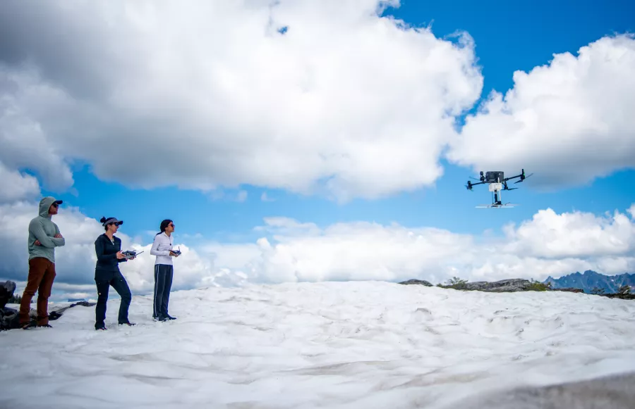 Three people stand on a glacier, one controlling a drone hovering nearby