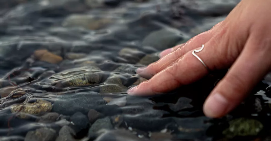 Caitlyn Blair's hand, touches a shallow rocky bottom of the water. On one of her fingers is a ring shaped like a wave. 