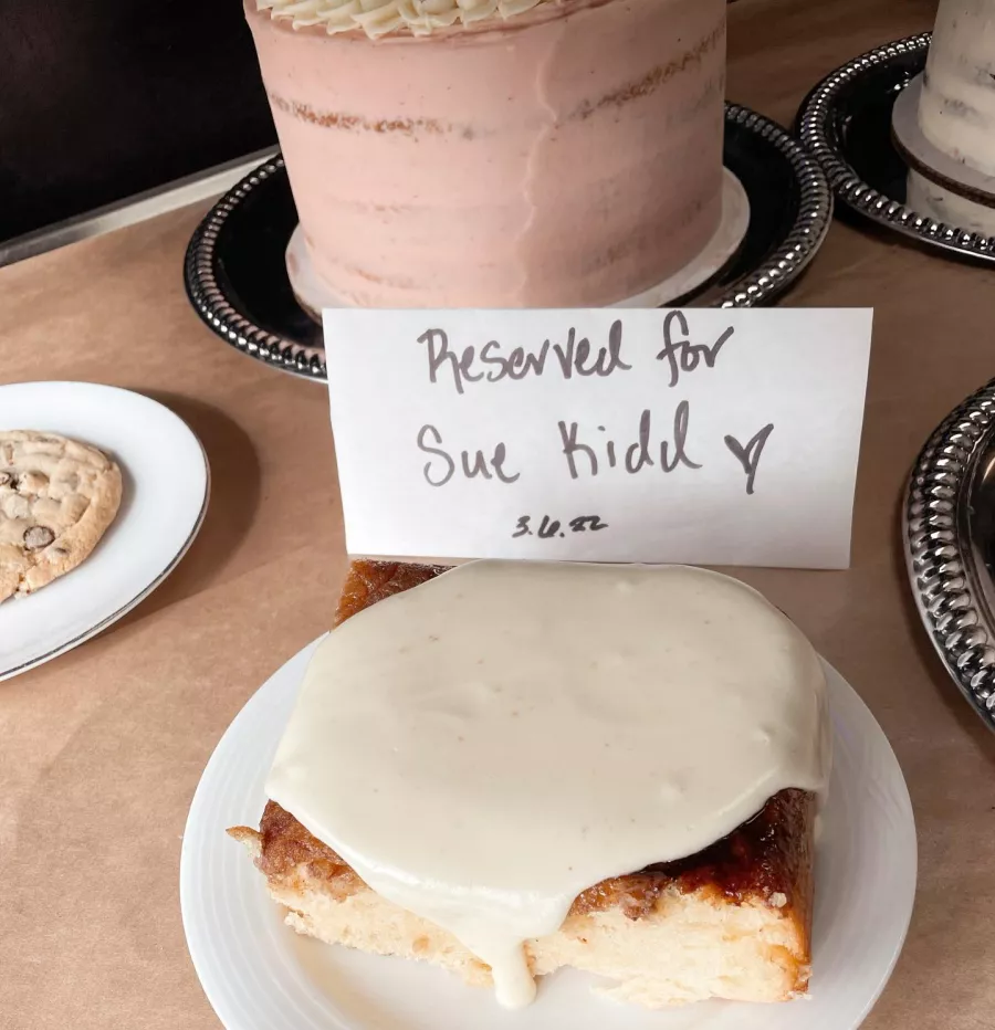 A huge cinnamon roll covered in thick white icing sits on a plate with a hand-printed sign, Reserved for Sue Kidd 3-6-22