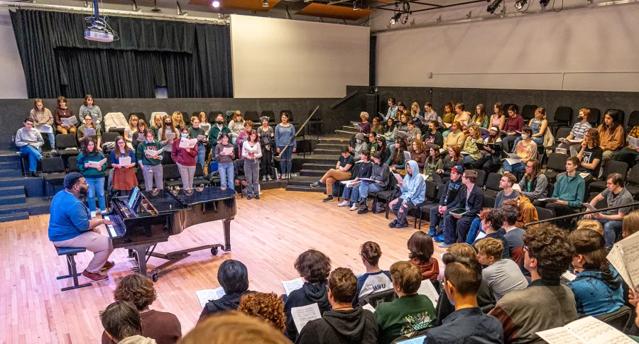 Hodges, at the piano, sits in the center of a rehearsal studio ringed with several rows of student choir members