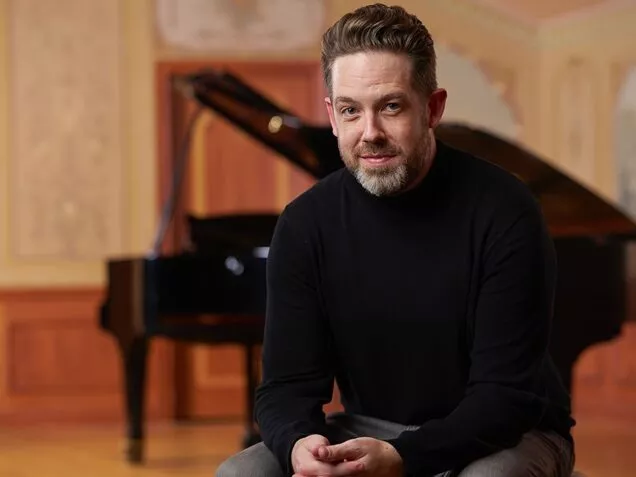 A man sits on a piano bench in front of a grand piano. He is wearing a black sweater and has short brown hair and a beard. 