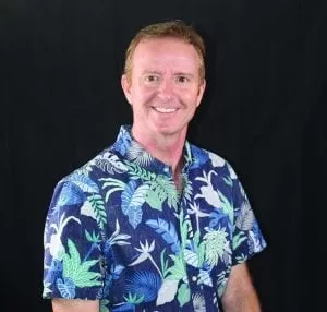 A man with short brown hair, wearing a blue Hawaiian shirt with green and white flowers, is smiling and looking directly at the camera. 