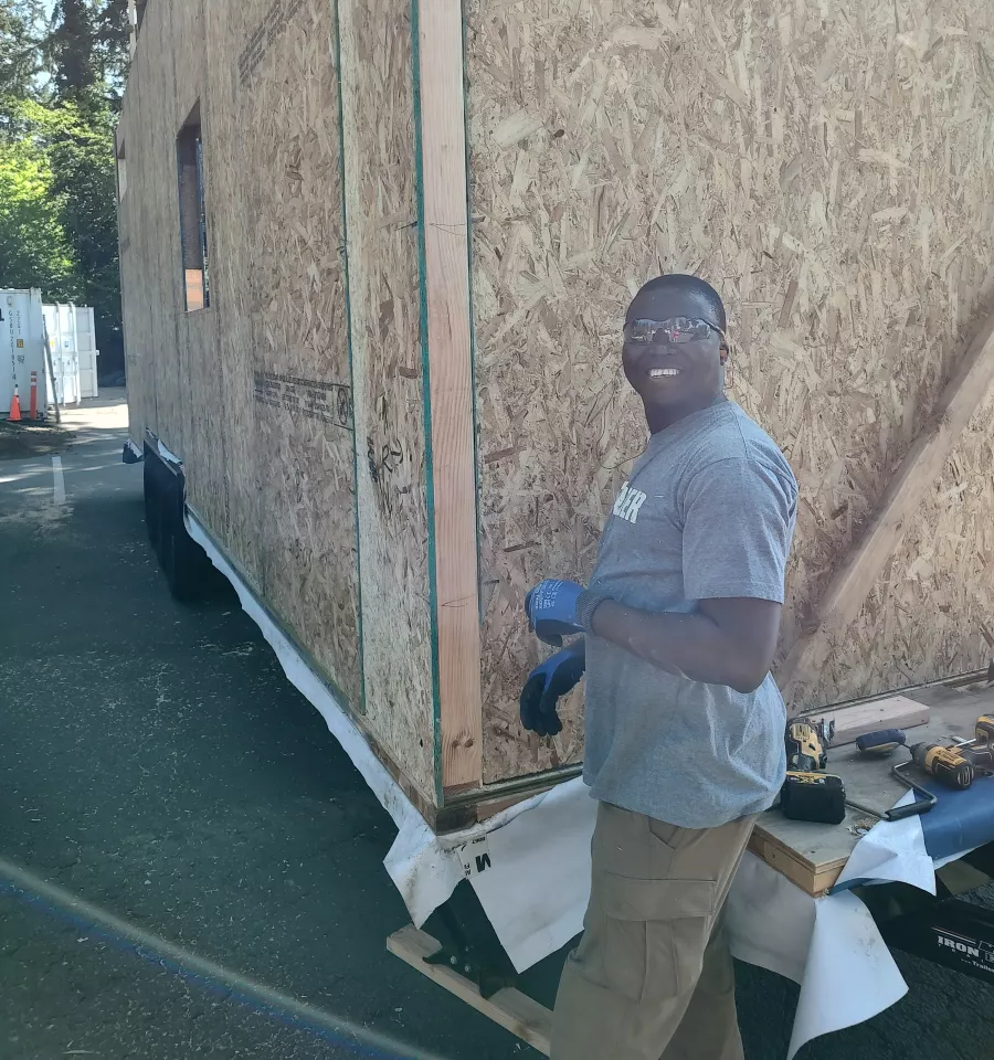 Singbo smiles next to an unfinished exterior wall at the tiny house construction project. 
