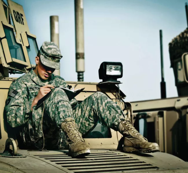 A soldier wearing fatigues sits on a tank, writing in a journal
