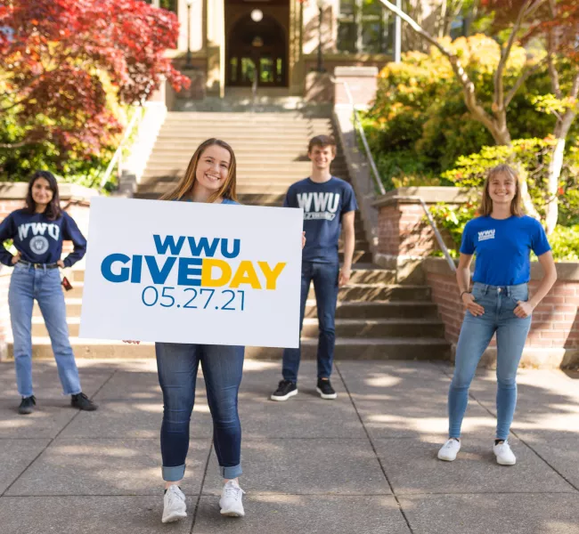 Four students wearing WWU t-shirts stand in front of Old Main; one holds a sign with WWU Give Day 05.27.21