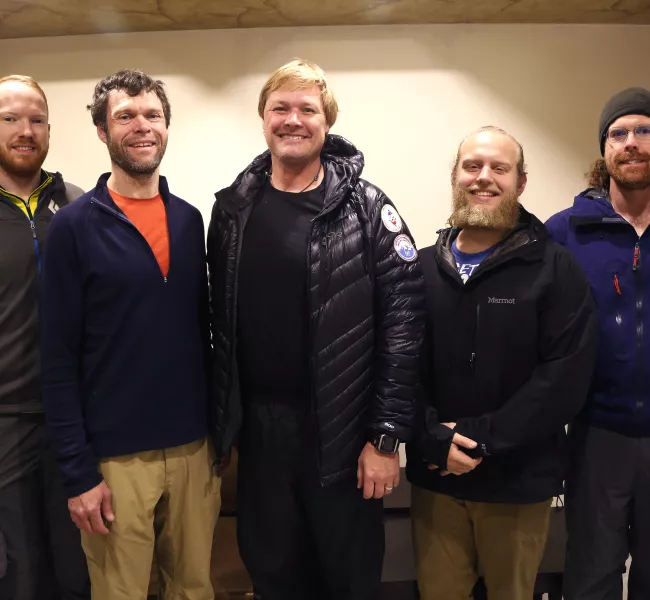 Five men wearing outdoor gear stand inside, smiling and facing the camera