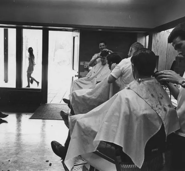 Three students sit in a line of barber chairs while barbers trim their hair. One student sits and waits. 