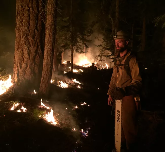 a fire fighter holds a chainsaw as he watches underbrush go up in flames