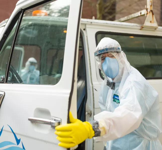 A man in full-body PPE opens the driver&#039;s-side door of a white van