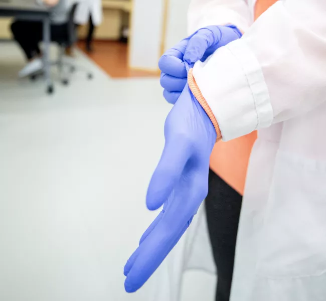 a student in a white lab coat dons a pair of blue plastic gloves