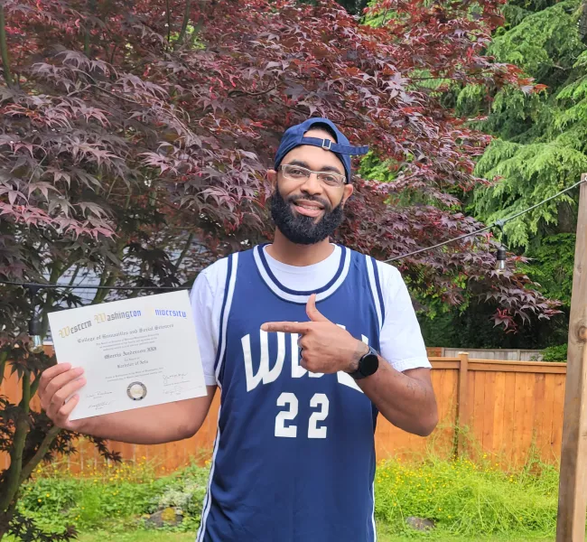 A person wearing a WWU basketball jersey smiles for the camera and points to a WWU diploma in his other hand. 
