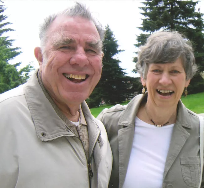 A man and a woman smile at the camera, surrounded by trees and lawn