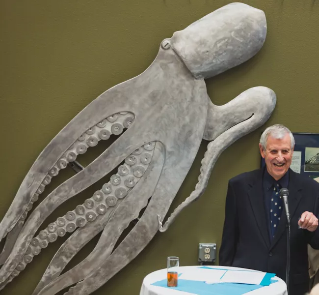 a person speaks at a microphone with a large sculpture of an octopus on the wall behind