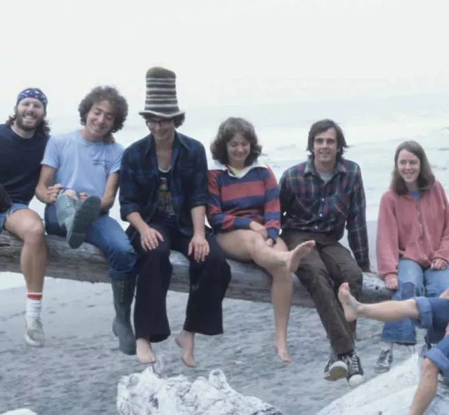 A 1970s photo of seven smiling students sitting on a driftwood log on the beach. One of the students is wearing a striped crocheted top hat. 