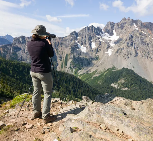 a woman uses a scope to scan the distant, craggy mountainside