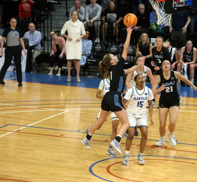 a woman reaches up for the basketball, surrounded by other players 