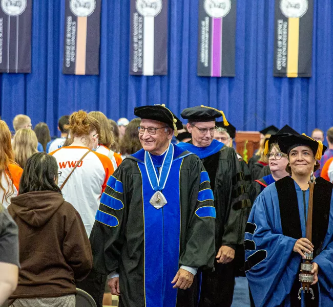 university administrators in academic regalia proceed through rows of standing students