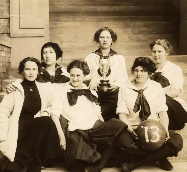 Six students sit on the steps of Old Main in 1913, displaying a trophy cup and a basketball.