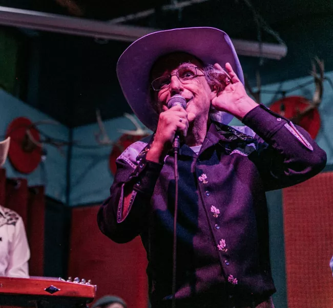 Patrick Haggerty, wearing a black cowboy shirt and lavender cowboy hat, sings on stage, holding one hand to his ear while he sings to encourage the audience to sing along. 