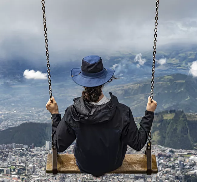 a student sits on a plank swing and swings out on a mountainside, a view of Quito far below them. 