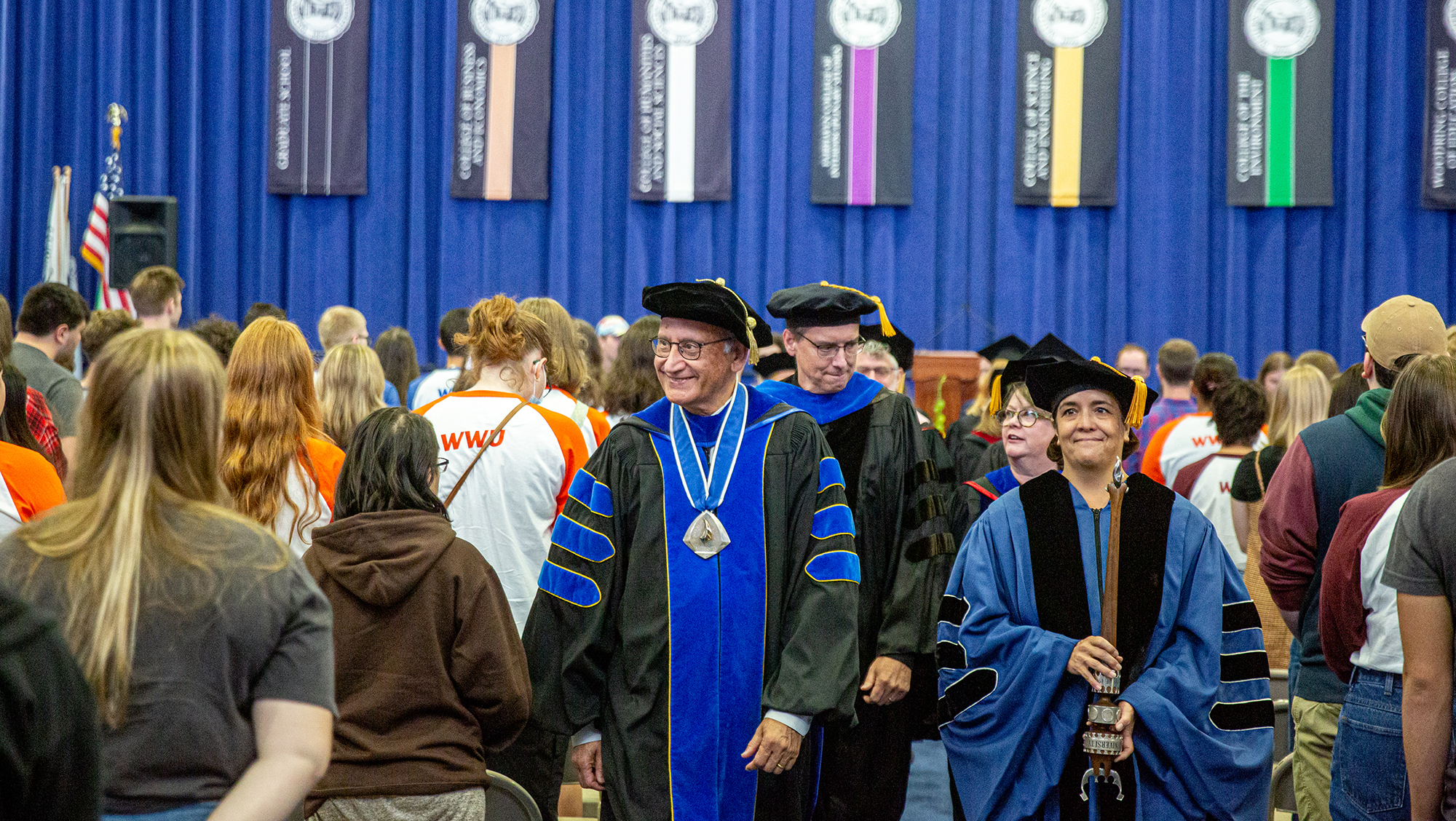 university administrators in academic regalia proceed through rows of standing students